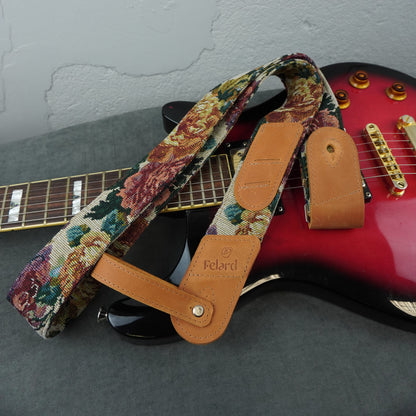 A guitar strap with a multicolor floral tapestry pattern and tan leather end pieces, laid across a red electric guitar on a gray surface against a textured gray wall. 