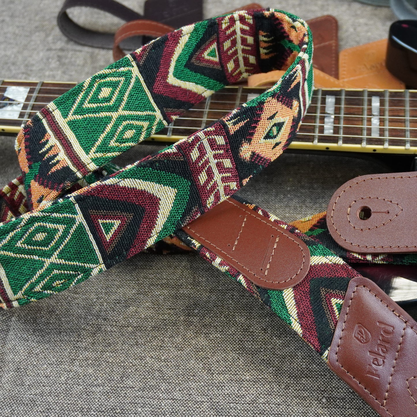 A close-up of a guitar strap with a colorful geometric pattern in green, red, beige, and black, featuring brown leather end pieces and a visible 'Iceland' brand patch.