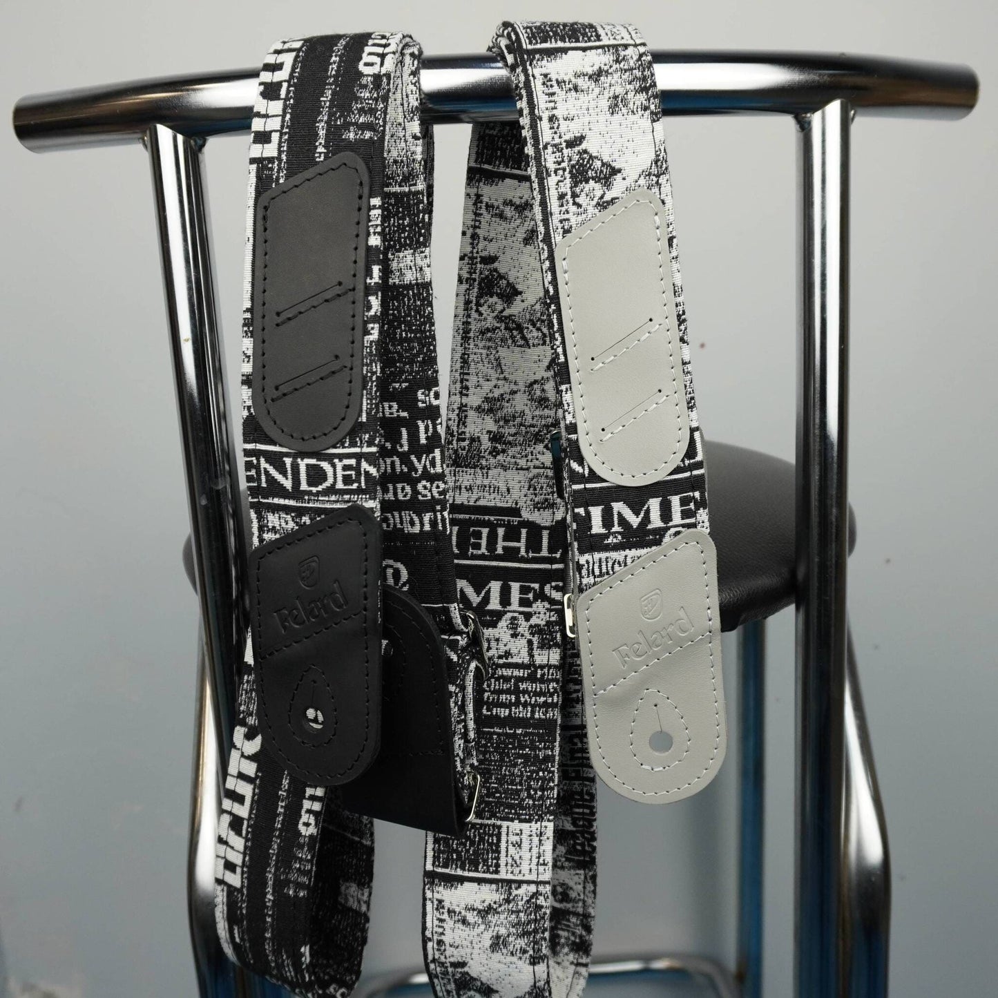 Two black and white newspaper-print shoulder straps with solid black and off-white leather patches, hanging from a chrome metal stand.