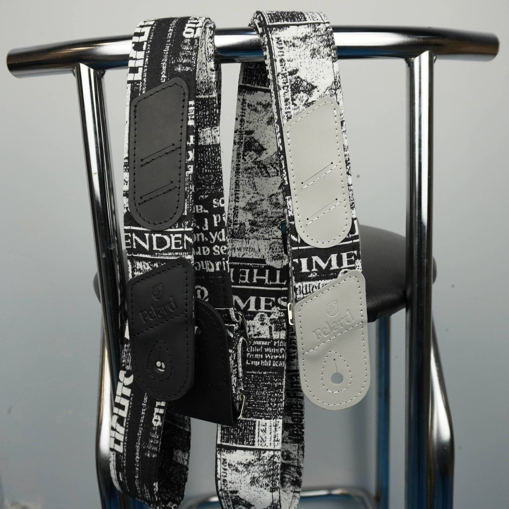 Two black and white newspaper-print shoulder straps with solid black and off-white leather patches, hanging from a chrome metal stand.