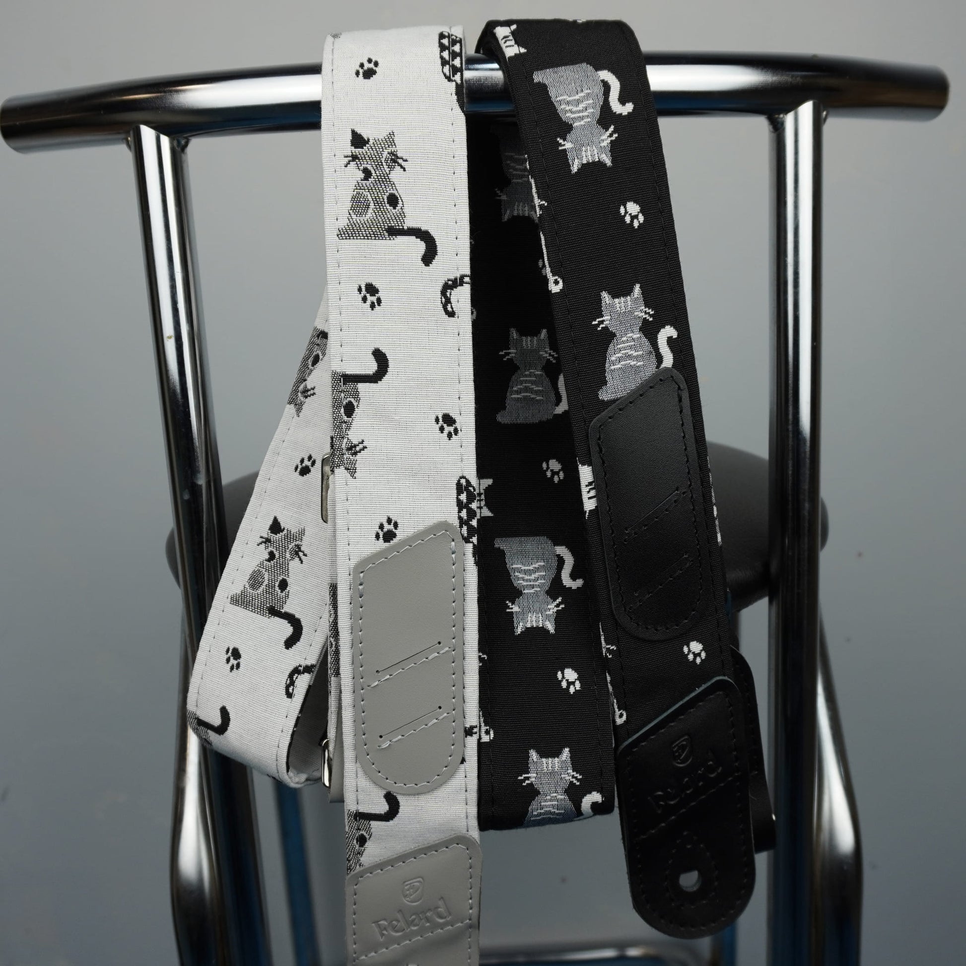 Two guitar straps hanging from a metal chair: one white with black cat and paw print patterns, and one black with gray cat and paw print patterns.