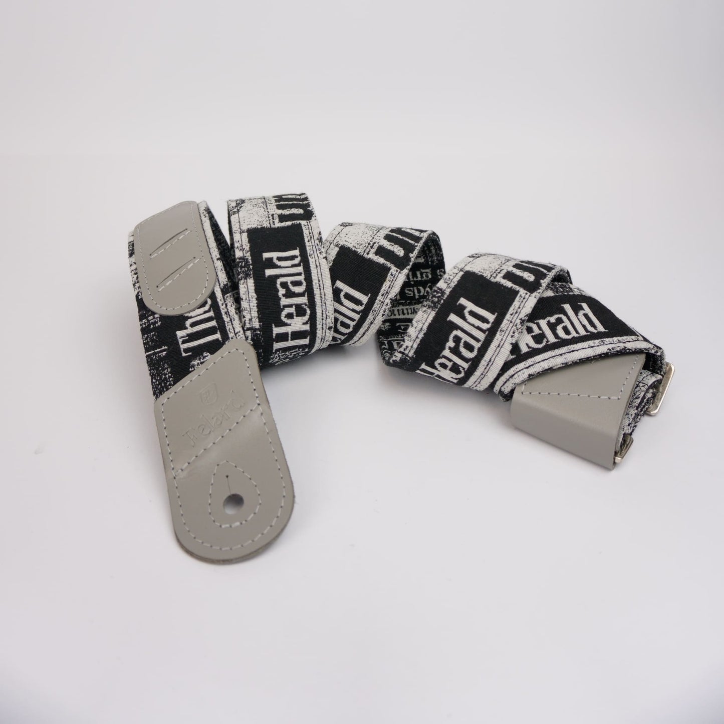 A guitar strap with a black and white newspaper print pattern featuring the text 'The Herald', finished with light gray leather ends and a silver metal buckle, displayed on a clean white background.