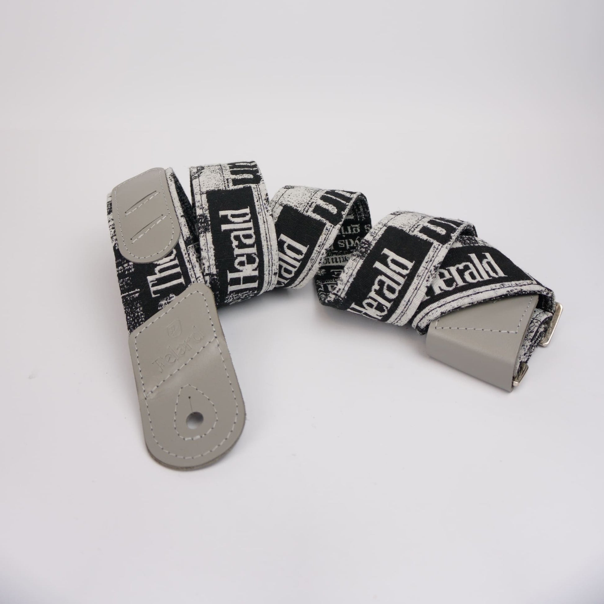 A guitar strap with a black and white newspaper print pattern featuring the text 'The Herald', finished with light gray leather ends and a silver metal buckle, displayed on a clean white background.