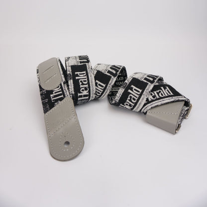 A guitar strap with a black and white newspaper print pattern featuring the text 'The Herald', finished with light gray leather ends and a silver metal buckle, displayed on a clean white background.