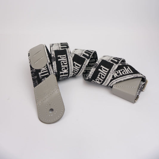 A guitar strap with a black and white newspaper print pattern featuring the text 'The Herald', finished with light gray leather ends and a silver metal buckle, displayed on a clean white background.