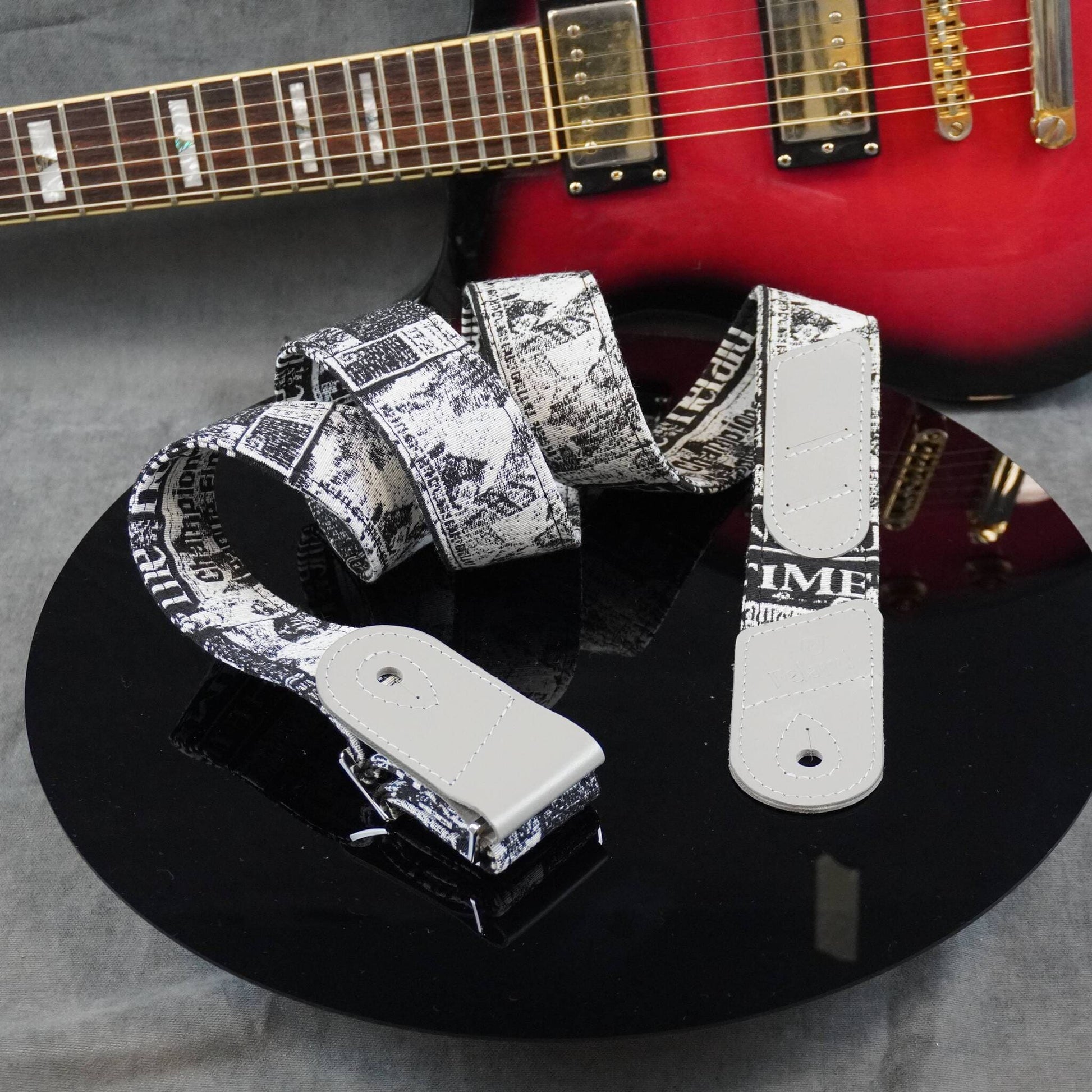 A guitar strap with a black and white newspaper print pattern and white metal hardware, displayed on a black reflective surface with a red electric guitar in the background.