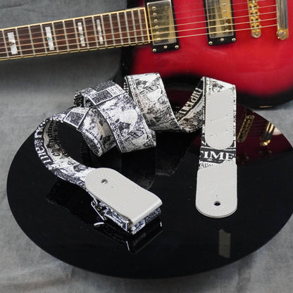 A guitar strap with a black and white newspaper print pattern and white metal hardware, displayed on a black reflective surface with a red electric guitar in the background.