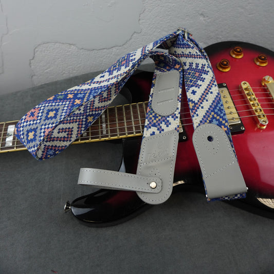 A guitar strap with a blue, white, and red geometric patterned fabric and light gray leather ends, draped over a red electric guitar on a gray surface.