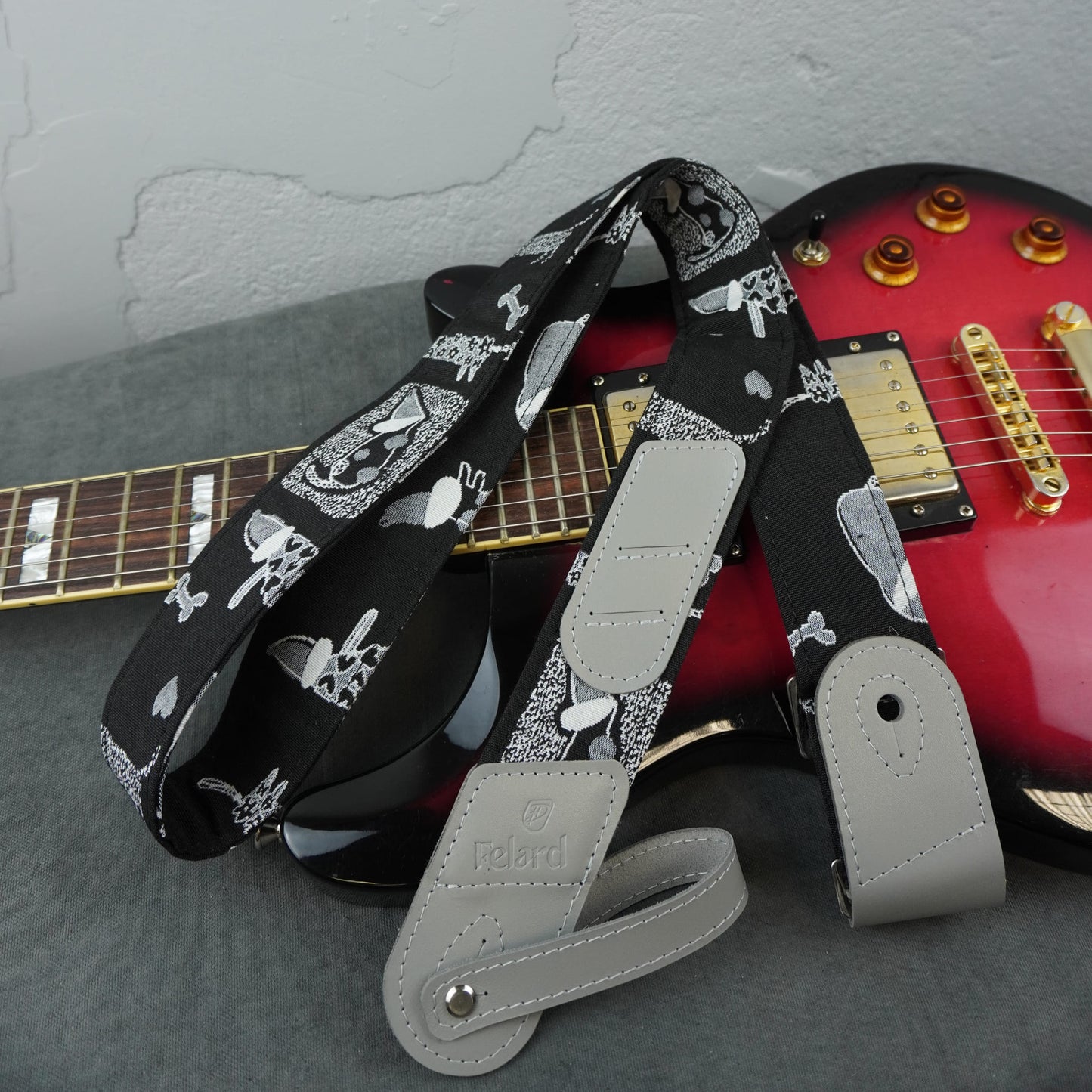 A black guitar strap with a white graphic print and light gray leather ends, laid over a red electric guitar on a gray surface against a textured gray wall.