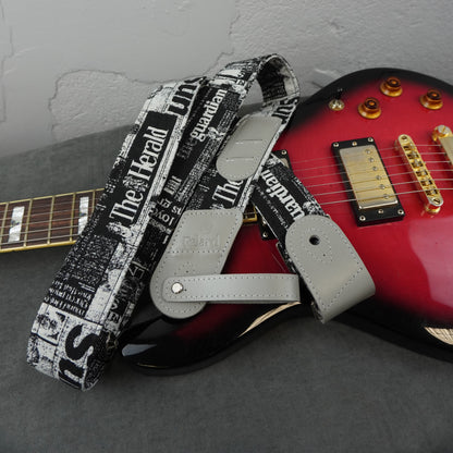 A guitar strap with a black and white newspaper print featuring 'The Herald' and 'The Guardian' text, laid across a red and black electric guitar on a gray surface.