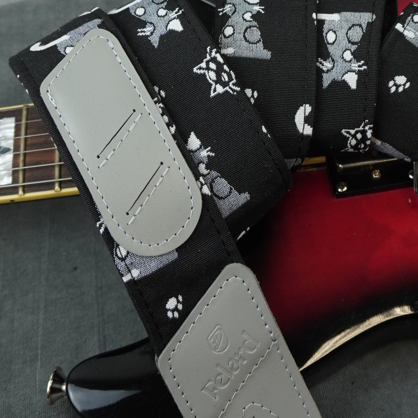 A close-up of a Felard electric guitar strap with a black and white cat print pattern and light gray padded shoulder straps, resting over a red and black guitar body.