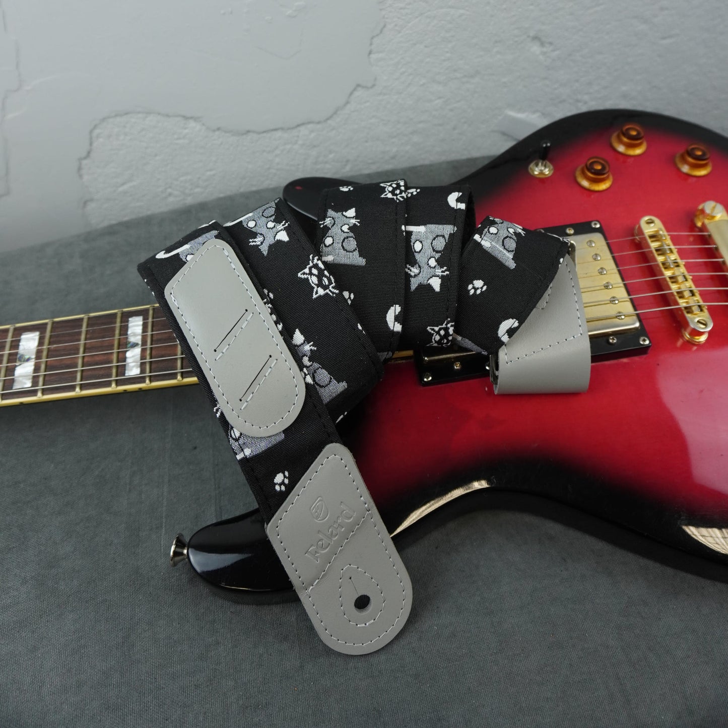A guitar strap with a black background and gray cat pattern, laid across a red electric guitar on a gray surface against a light gray wall.