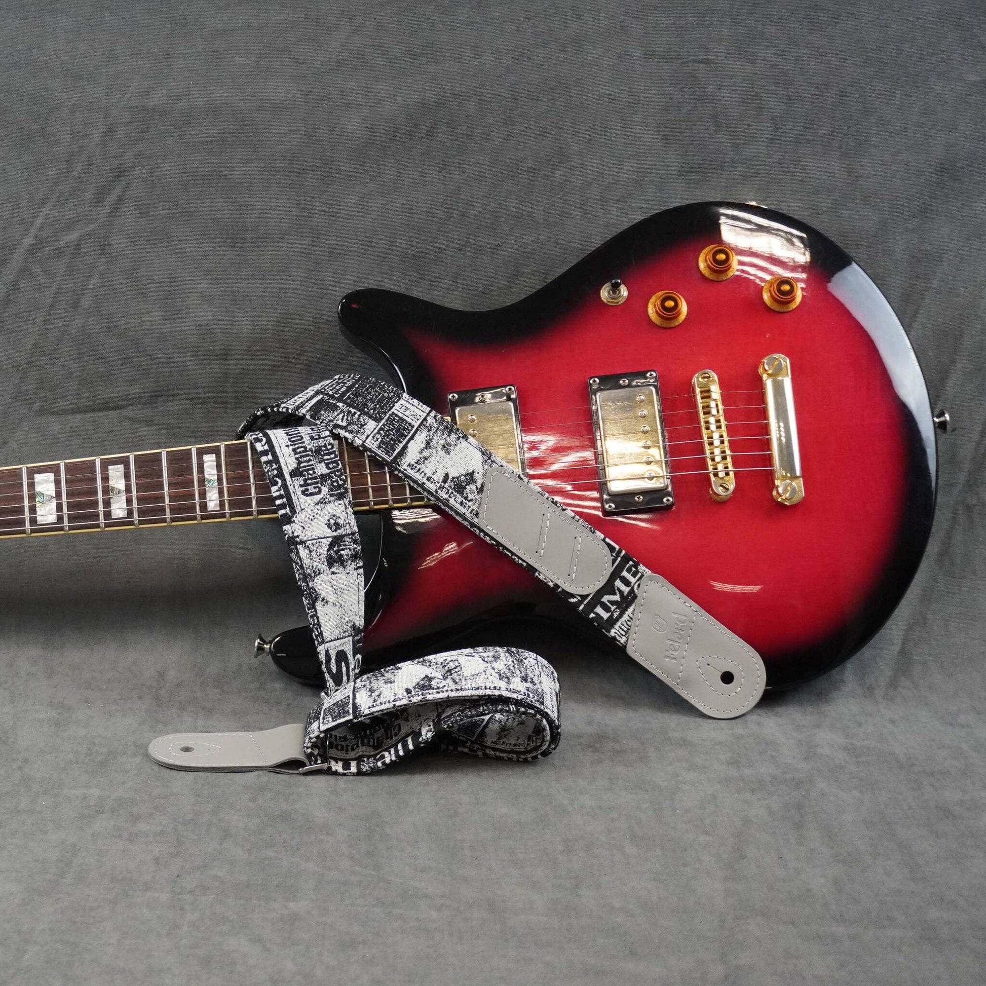 A red and black electric guitar with a black-and-white patterned guitar strap, photographed on a gray fabric background.