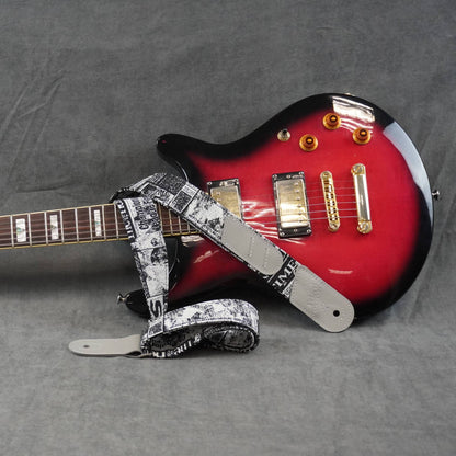 A red and black electric guitar with a black-and-white patterned guitar strap, photographed on a gray fabric background.