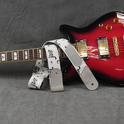 A guitar strap with a white background and black cat print pattern, draped over the neck of a red electric guitar on a gray textured background.