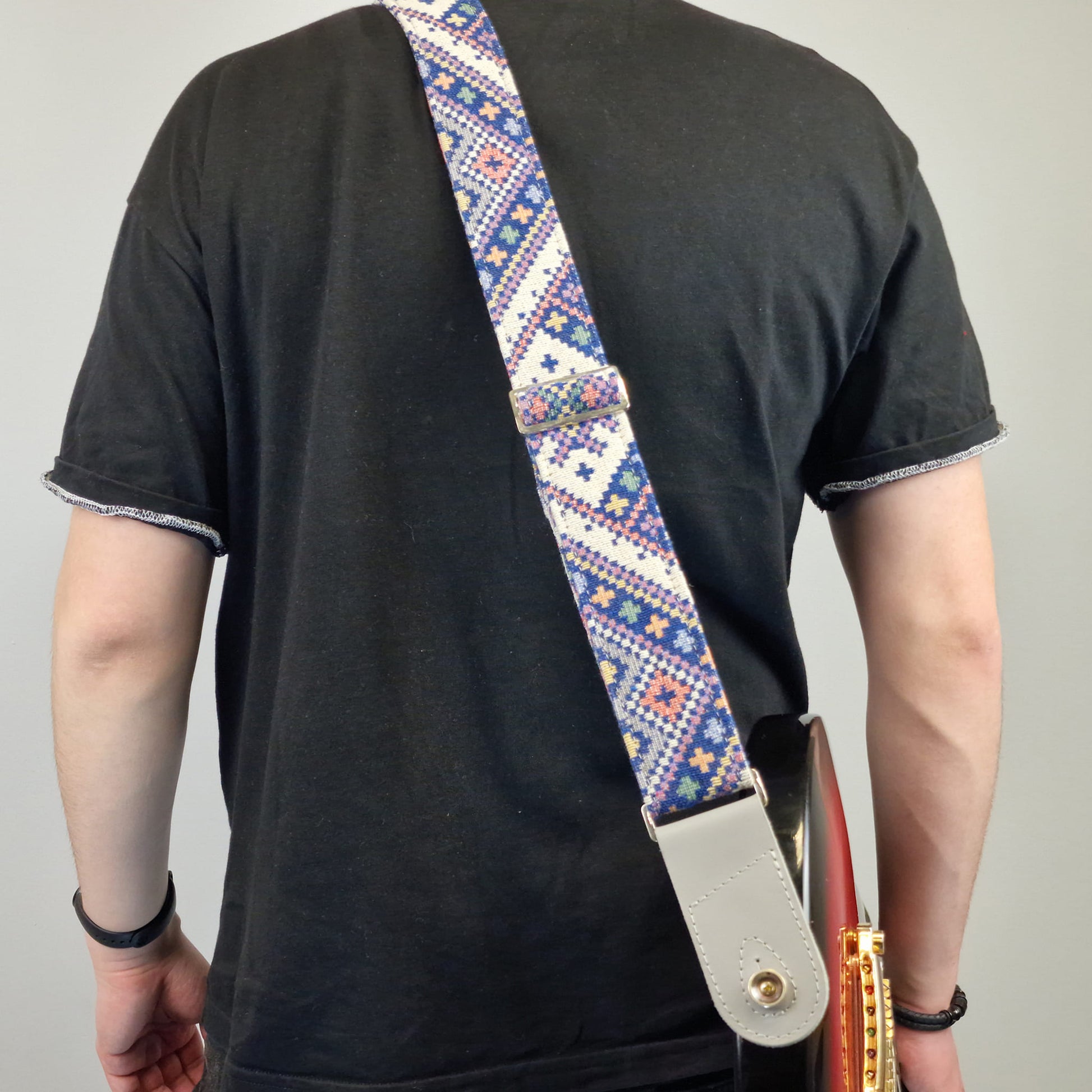  decorative guitar strap. The strap features a white base with a colorful geometric pattern in blue, red, and yellow, and has a light gray leather end piece.