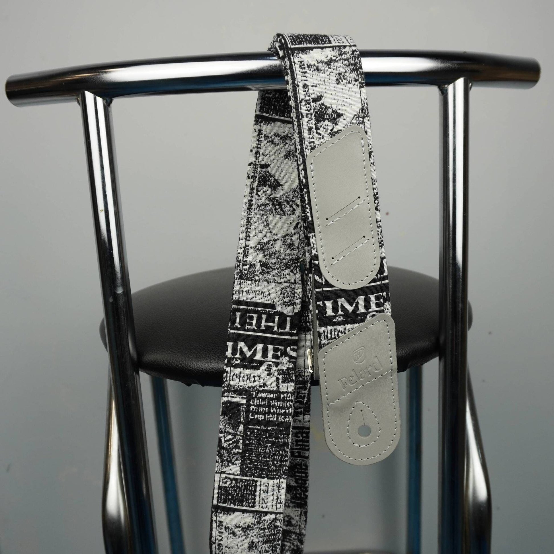 A guitar strap with a black and white newspaper print pattern and light beige leather ends, hanging from the backrest of a chrome metal chair.