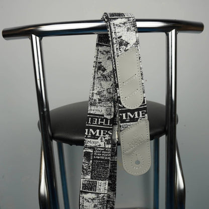 A guitar strap with a black and white newspaper print pattern and light beige leather ends, hanging from the backrest of a chrome metal chair.