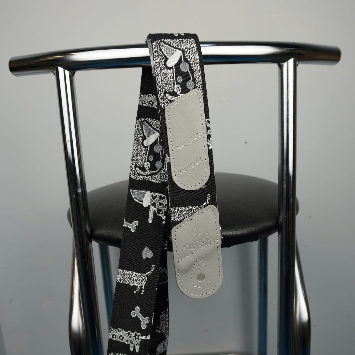 A black guitar strap with a white and gray dog bandana print and light gray adjustable plastic sliders, draped over the back of a chrome metal chair.