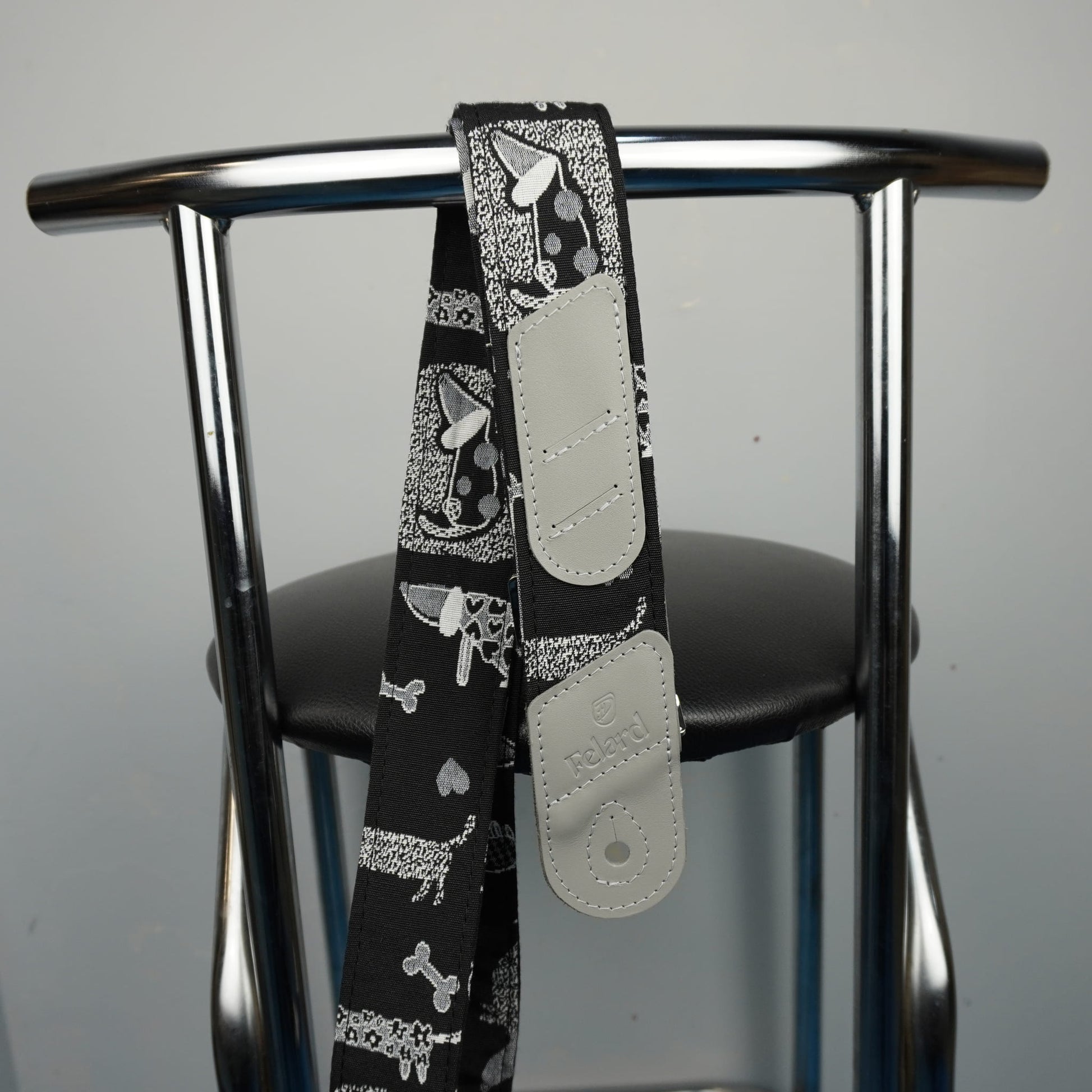 A black guitar strap with a white and gray dog bandana print and light gray adjustable plastic sliders, draped over the back of a chrome metal chair.