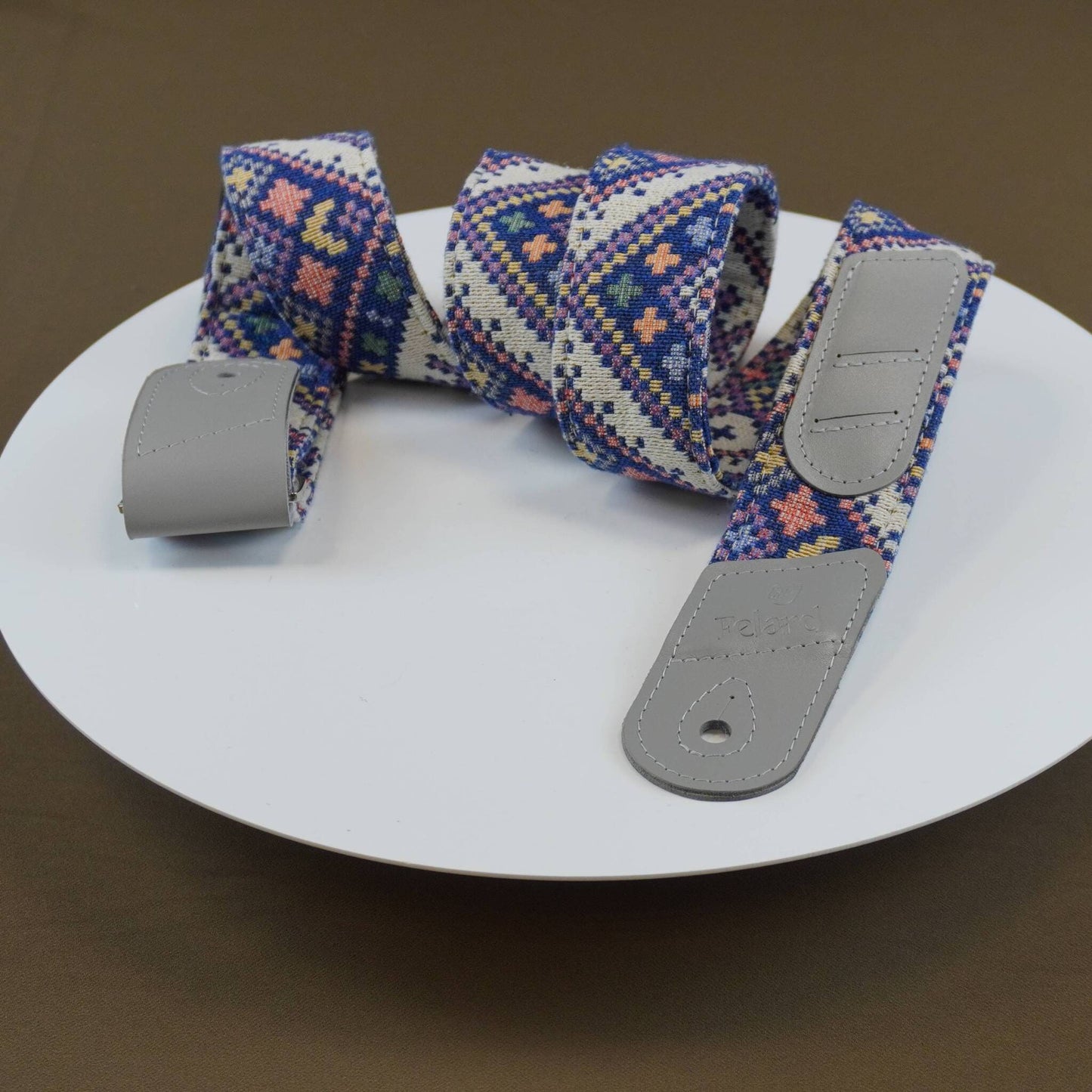A woven fabric guitar strap with a geometric pattern in blue, pink, and white, featuring gray leather-like end caps, displayed on a white round plate.