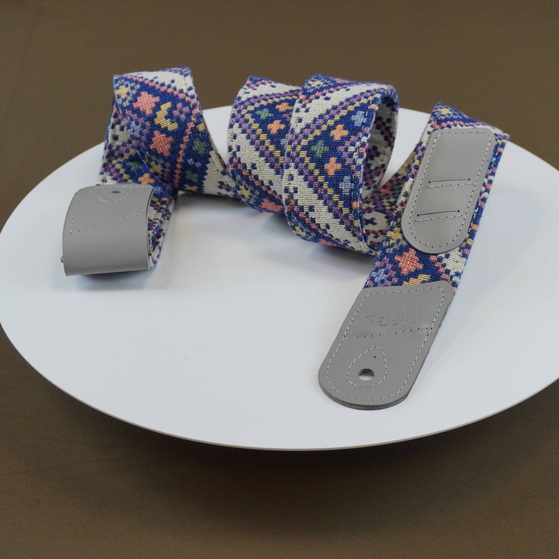 A woven fabric guitar strap with a geometric pattern in blue, pink, and white, featuring gray leather-like end caps, displayed on a white round plate.