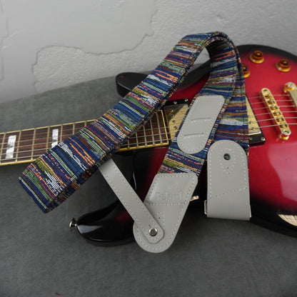 A guitar strap with a colorful striped woven pattern and light gray leather ends, draped over a red electric guitar on a gray surface against a white wall.