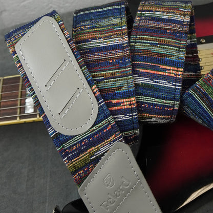 A close-up of a Felard guitar strap with a multicolor striped woven fabric and light gray leather end pieces, draped over part of a guitar.