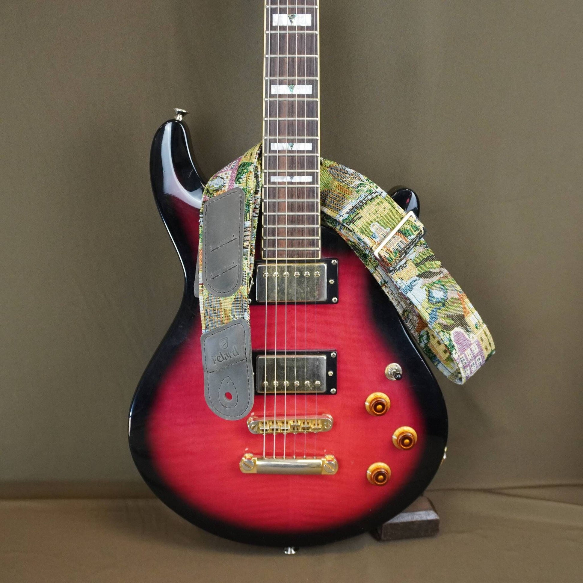 A custom electric guitar with a red-to-black sunburst finish, featuring a colorful floral patterned strap and gray pickups, displayed on a stand against a solid brown background.