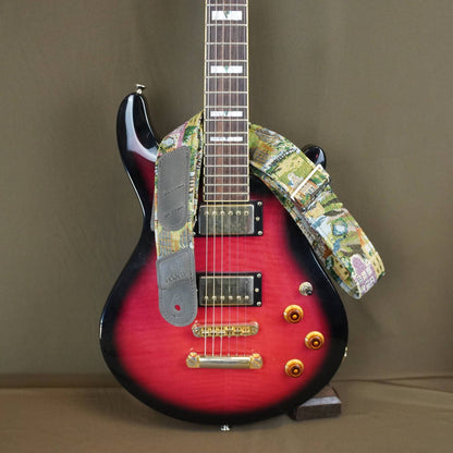 A custom electric guitar with a red-to-black sunburst finish, featuring a colorful floral patterned strap and gray pickups, displayed on a stand against a solid brown background.