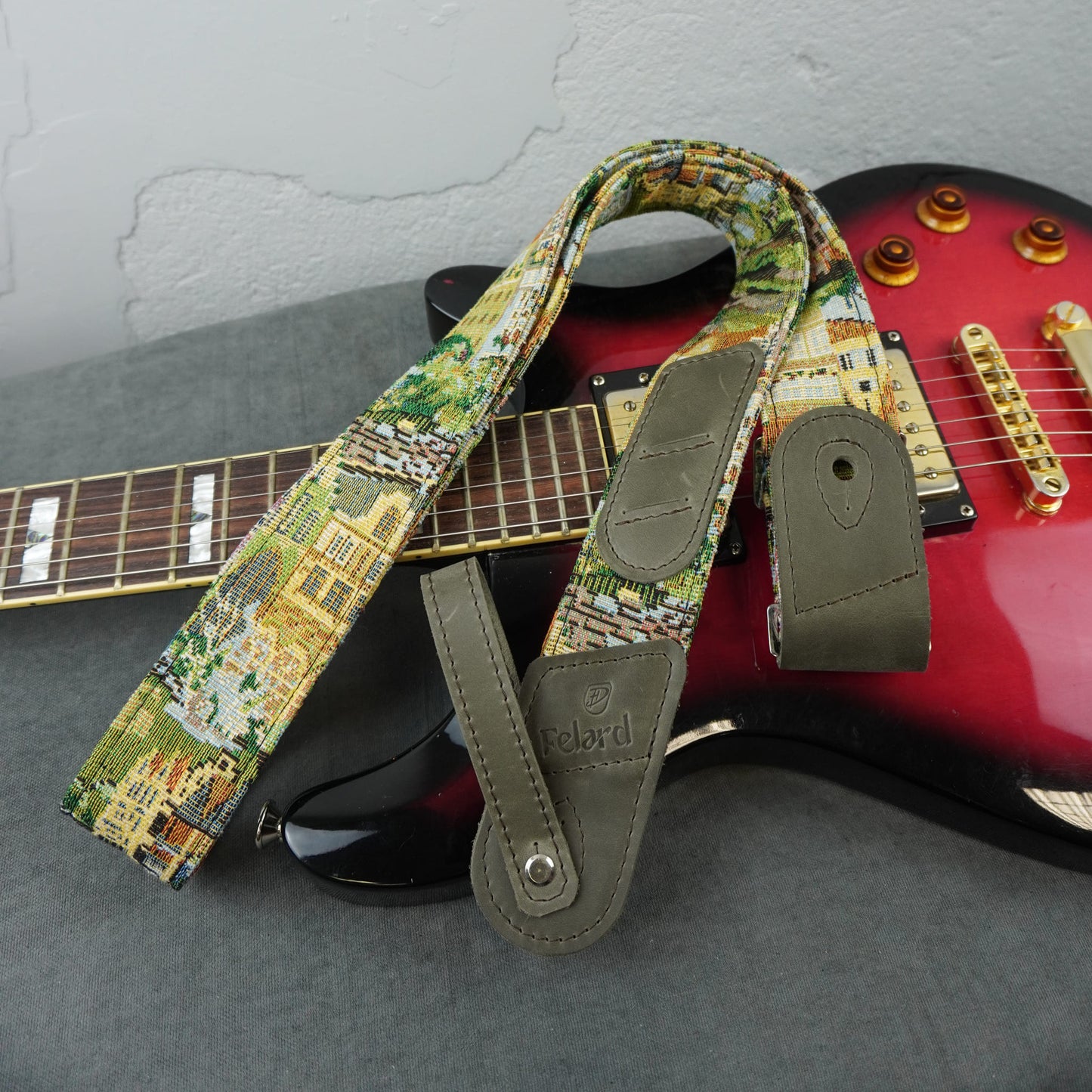 A guitar strap with a colorful scenic print and olive green leather ends, draped over a red electric guitar on a gray surface.