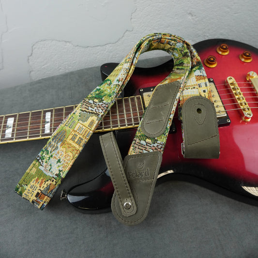 A guitar strap with a colorful scenic print and olive green leather ends, draped over a red electric guitar on a gray surface.