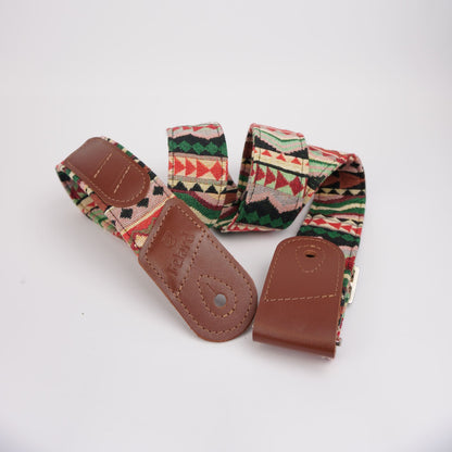 A vintage guitar strap with a colorful geometric patterned fabric and brown leather ends, displayed on a clean white background.