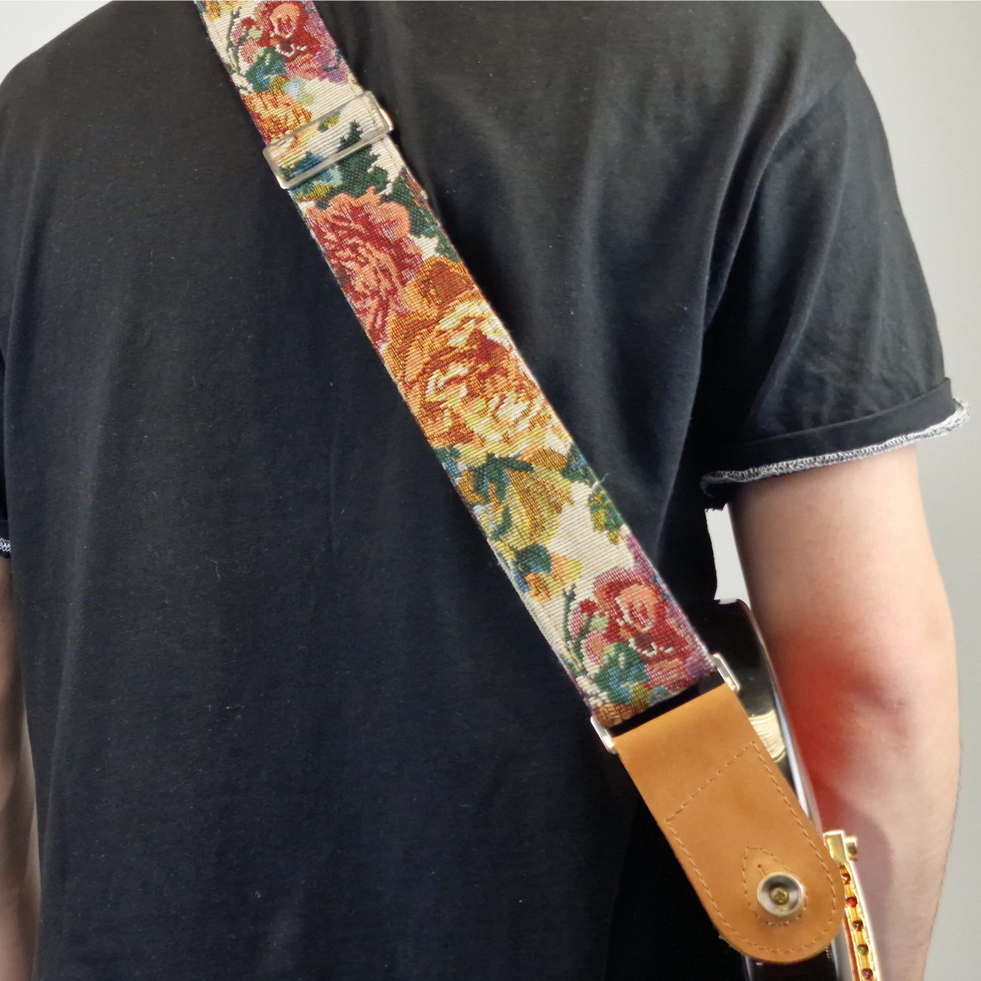 A close-up of a person  playing an electric guitar, showing a detailed view of a floral patterned guitar strap with a tan leather end piece.
