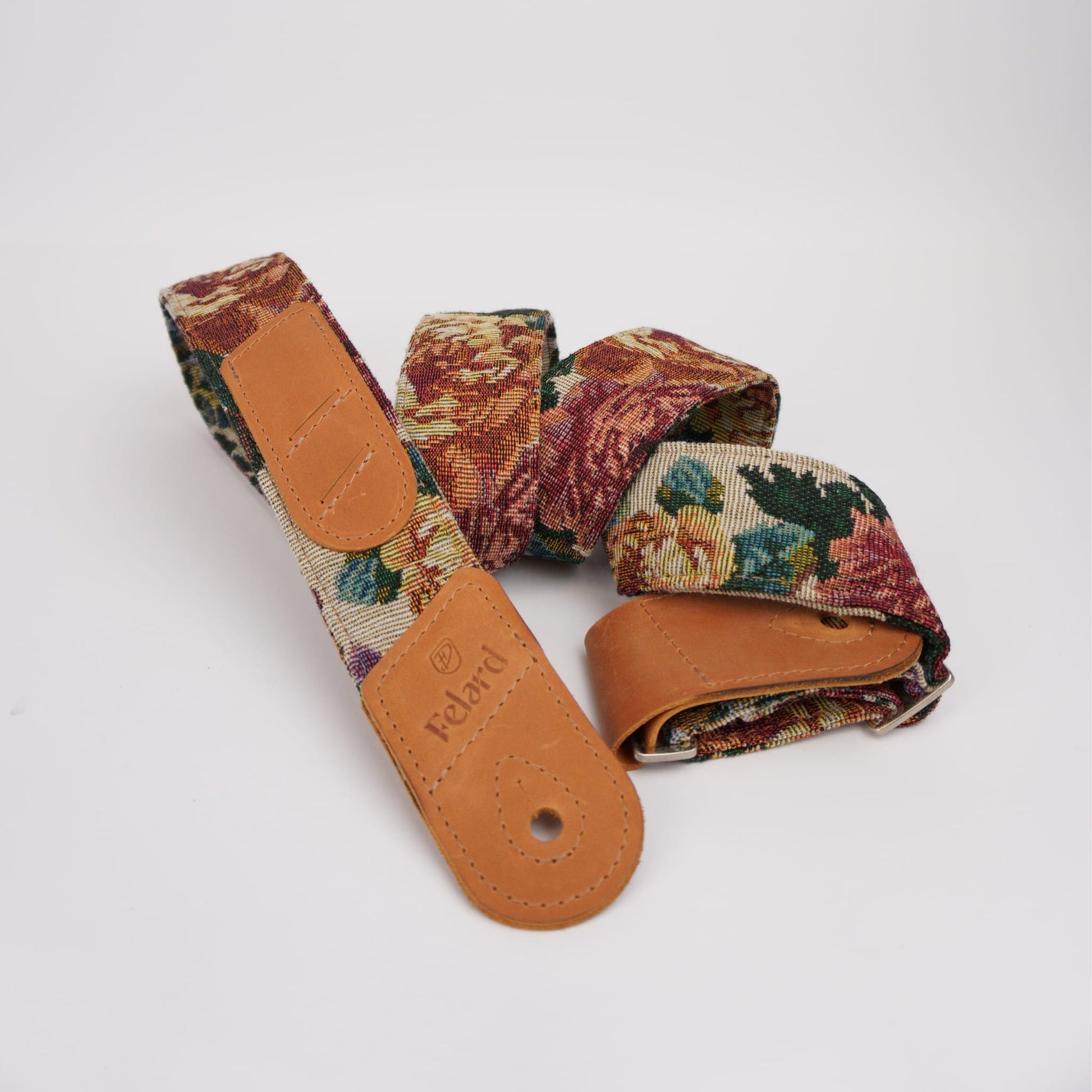 A vintage guitar strap with a multicolor floral tapestry pattern and tan leather ends, featuring the embossed brand name 'Felard' on one end, displayed on a clean white background.