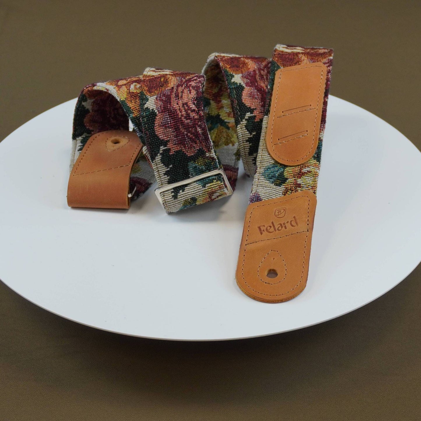 A floral tapestry-style guitar strap with tan leather ends, displayed on a white plate against a dark brown background. The brand name 'Felard' is visible on one of the leather tabs.