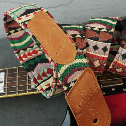 A close-up of a guitar strap with a colorful geometric woven pattern and tan leather end pieces, resting on a guitar neck. The brand name 'Felard' is embossed on one leather tab.