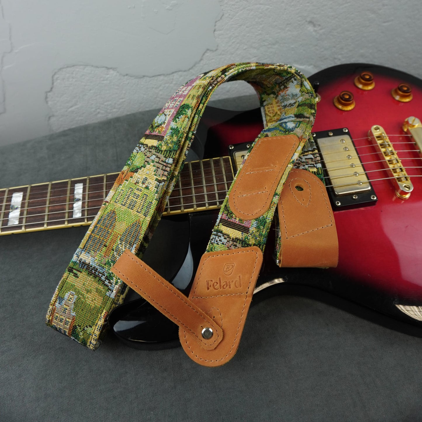 A guitar strap with a colorful landscape print and tan leather ends, laid across a red electric guitar on a gray surface.