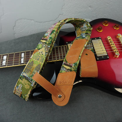A guitar strap with a colorful landscape print and tan leather ends, laid across a red electric guitar on a gray surface.