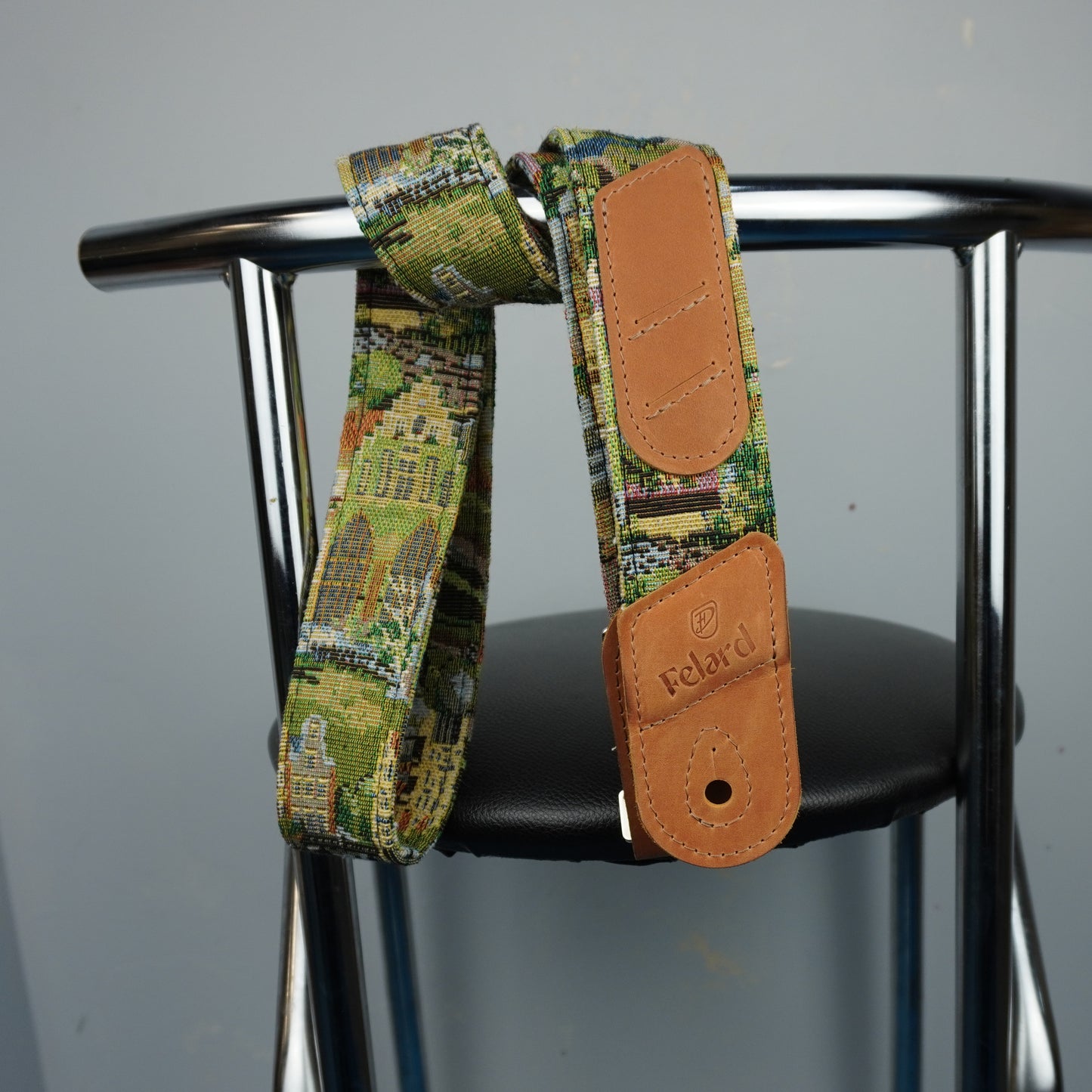 A close-up of a guitar strap with a green and yellow architectural pattern and tan leather end pieces, hanging from a black chair seat against a gray wall. The brand name 'Felard is visible on the leather end cap.