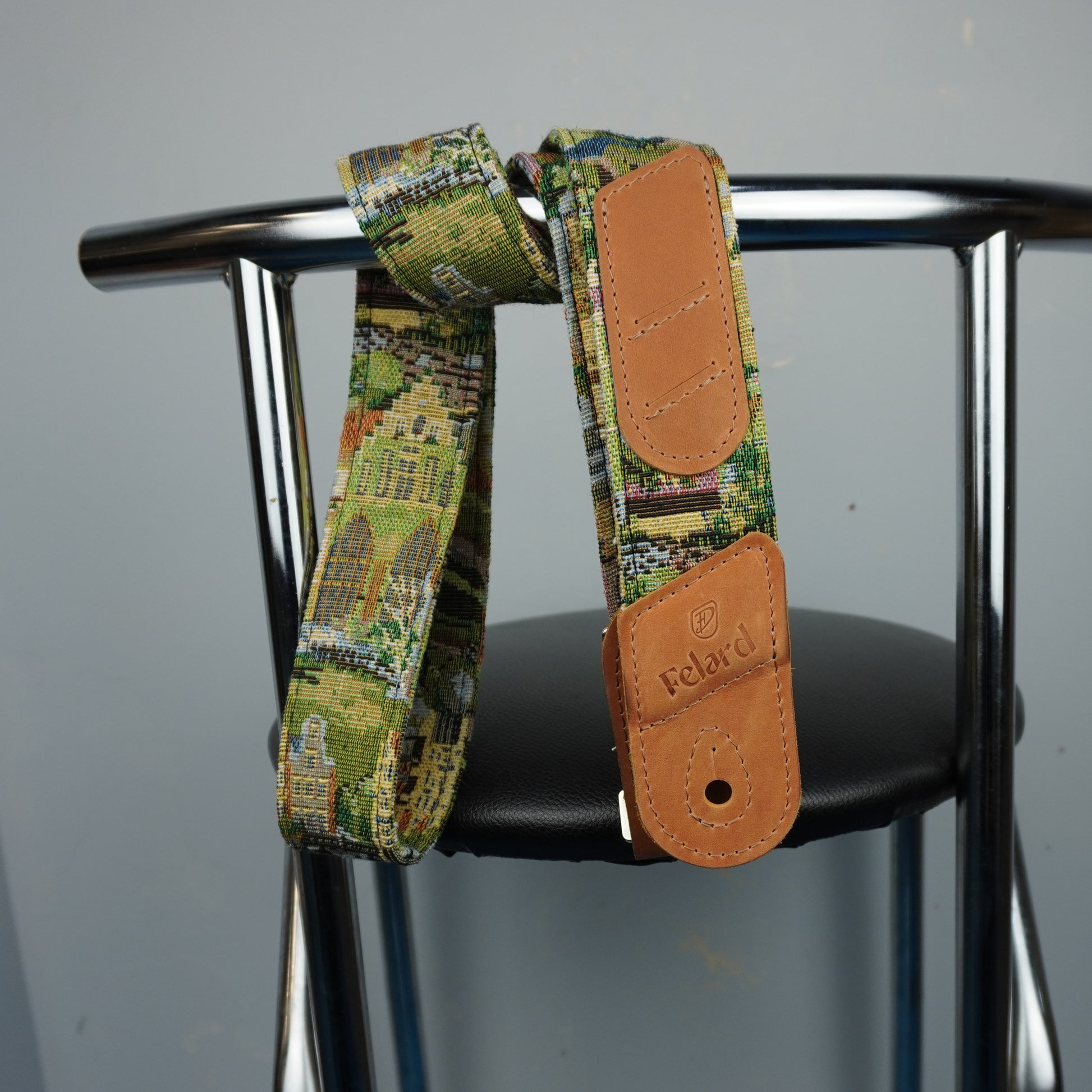 A close-up of a guitar strap with a green and yellow architectural pattern and tan leather end pieces, hanging from a black chair seat against a gray wall. The brand name 'Felard is visible on the leather end cap.