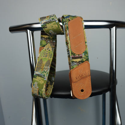 A close-up of a guitar strap with a green and yellow architectural pattern and tan leather end pieces, hanging from a black chair seat against a gray wall. The brand name 'Felard is visible on the leather end cap.