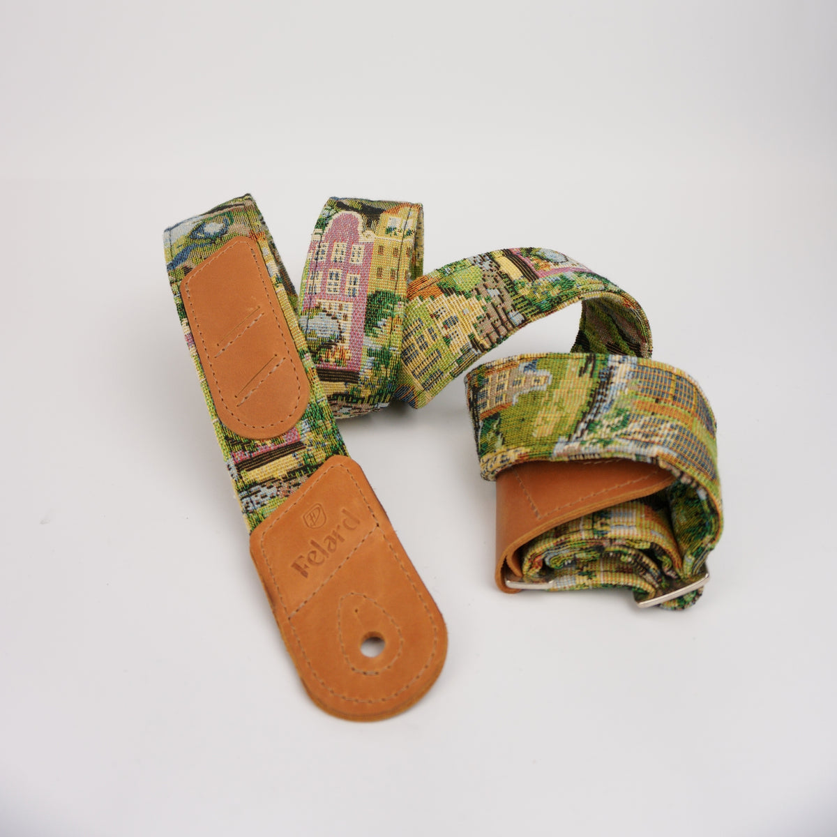 A guitar strap with a colorful, vintage-style town scene print in green, yellow, and pink, featuring tan leather end caps. The brand name 'Felard' is embossed on one of the leather sections.