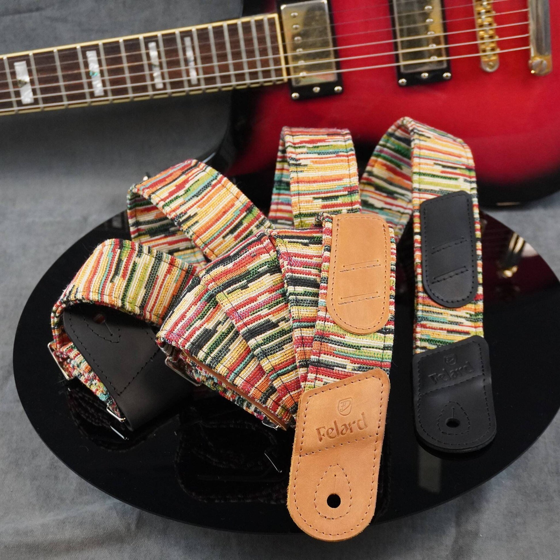 Guitar strap with a colorful striped fabric body and tan leather end pieces, displayed on a black reflective surface with a red electric guitar blurred in the background.