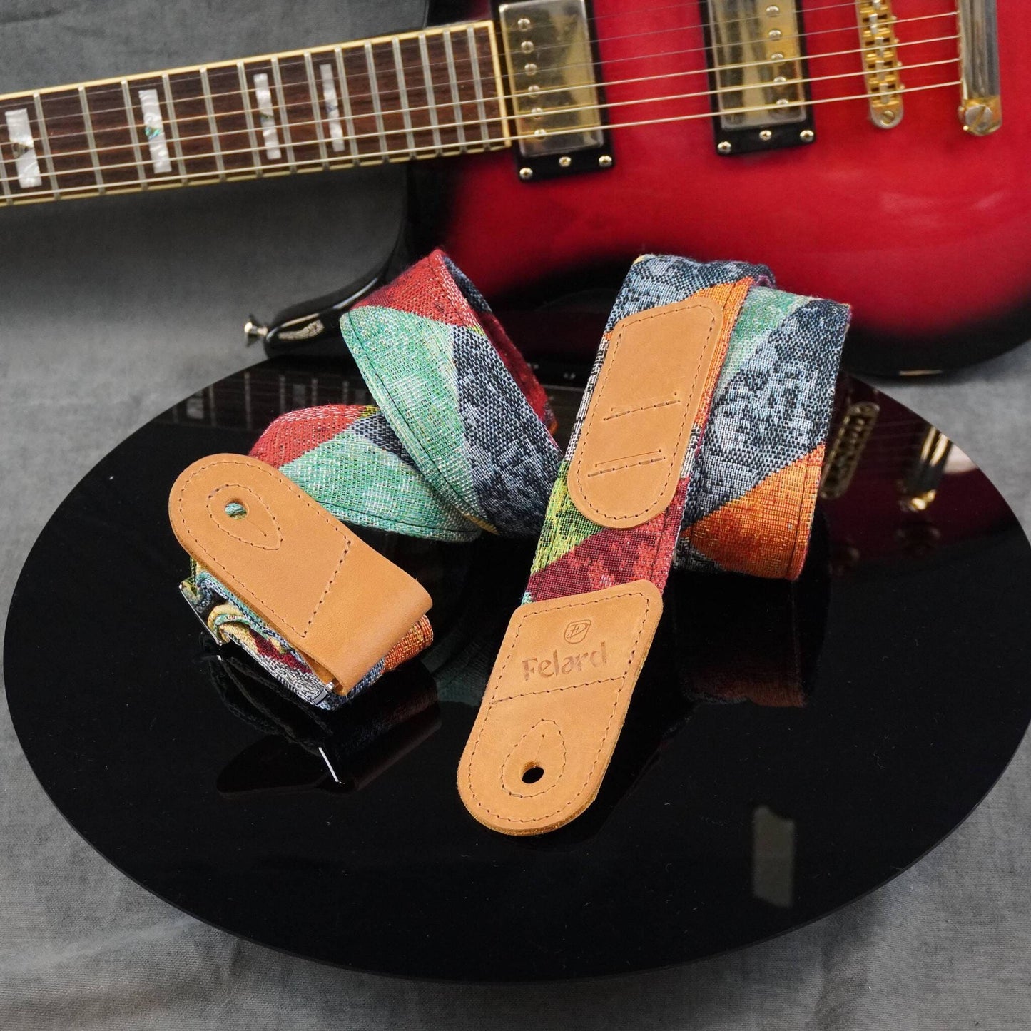 Felard guitar strap with a multicolor patchwork pattern and tan leather ends, displayed on a black reflective surface with a red electric guitar in the background.