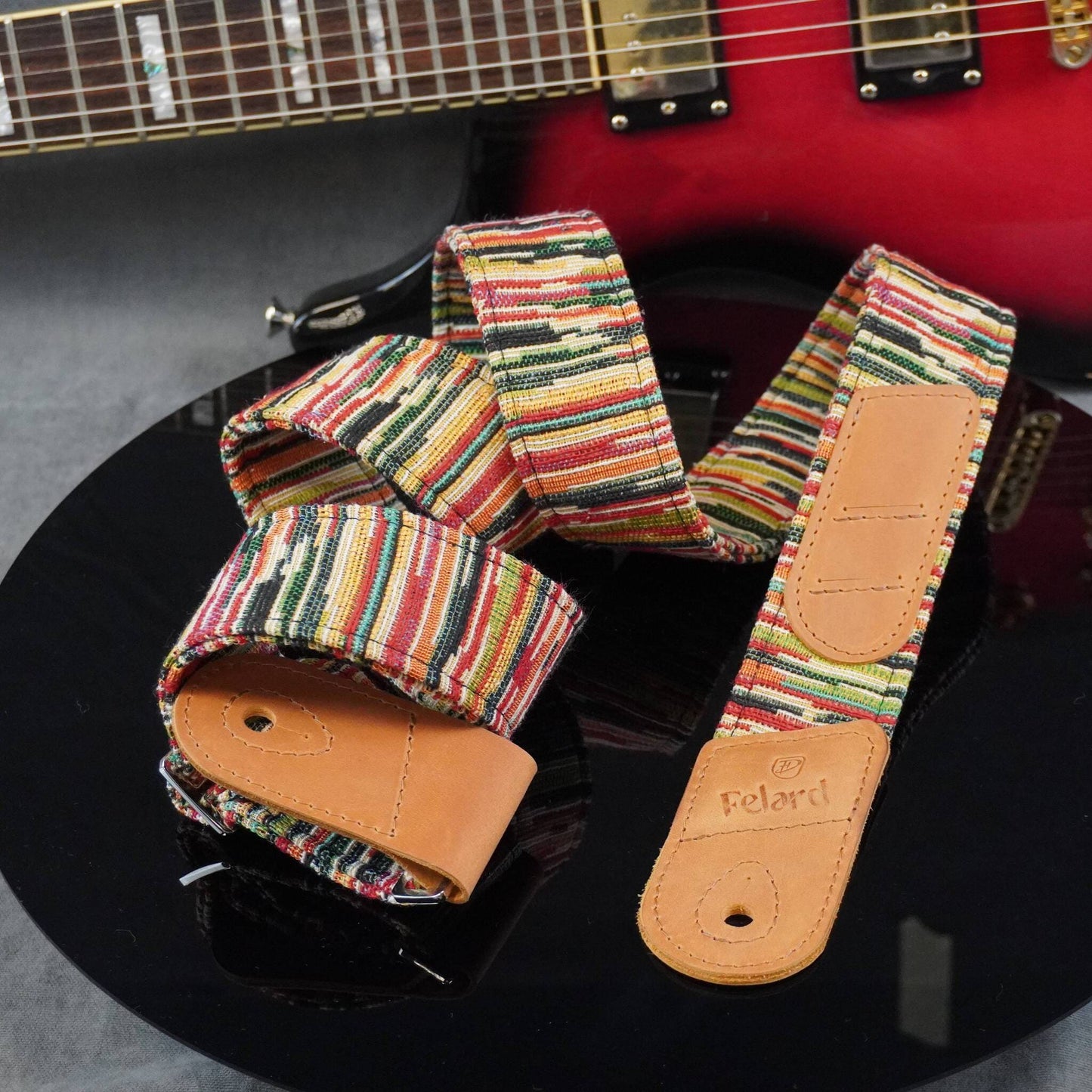 multicolor striped guitar strap with tan leather end pieces, displayed on a black reflective surface with a red electric guitar partially visible in the background.