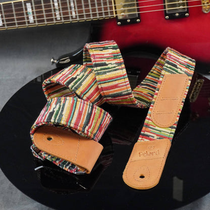 multicolor striped guitar strap with tan leather end pieces, displayed on a black reflective surface with a red electric guitar partially visible in the background.