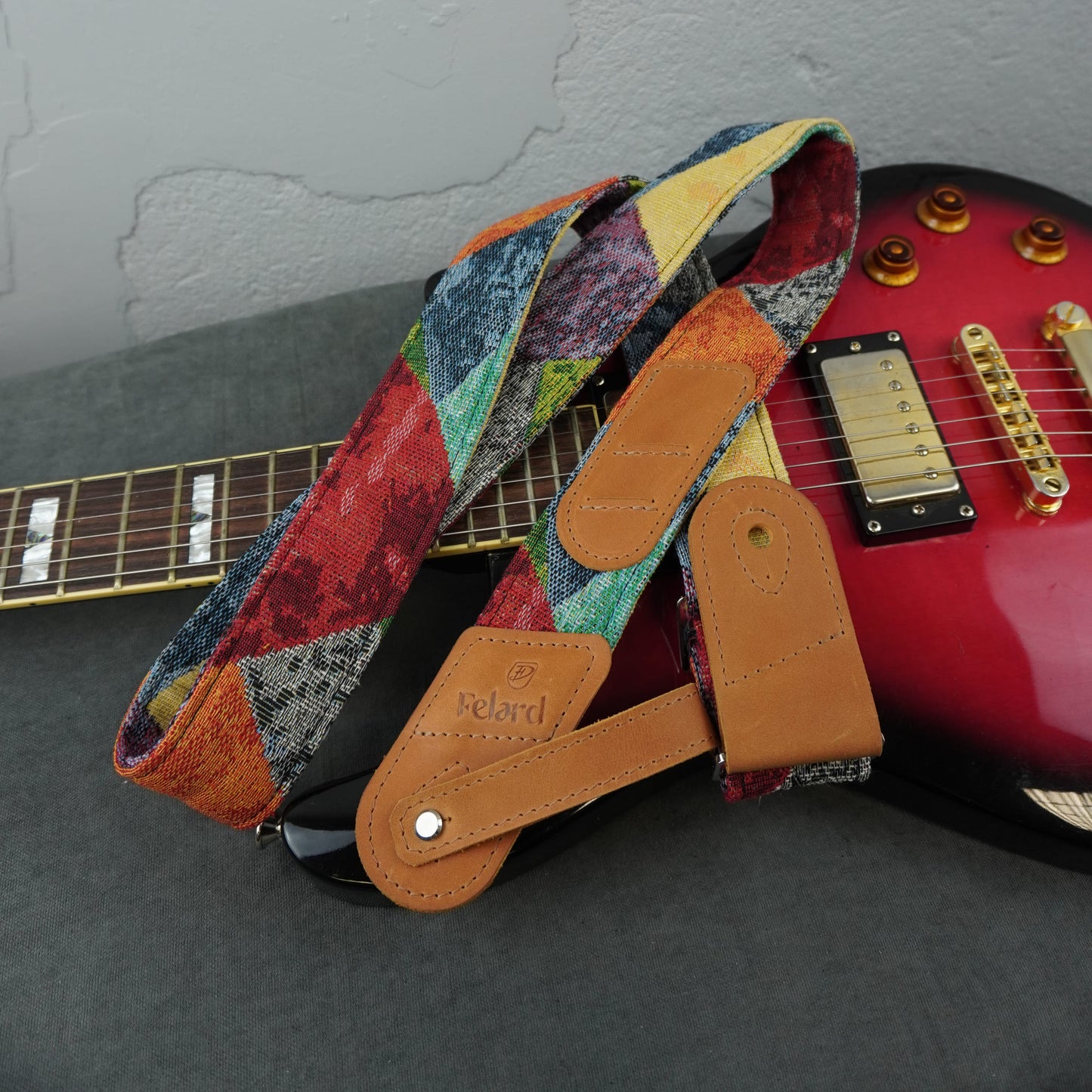 A multicolor patchwork-patterned guitar strap with tan leather end pieces and a silver metal snap, draped over a red electric guitar on a dark gray surface.