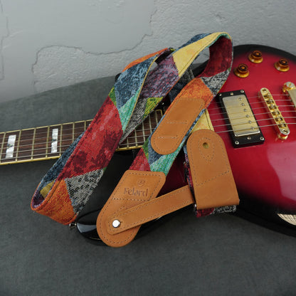 A multicolor patchwork-patterned guitar strap with tan leather end pieces and a silver metal snap, draped over a red electric guitar on a dark gray surface.