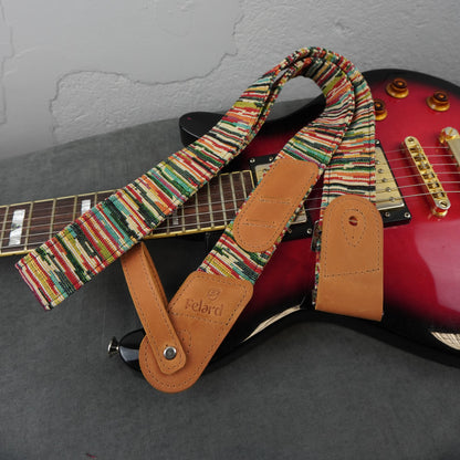 A multicolor striped fabric guitar strap with tan leather ends, shown draped over a red electric guitar on a gray surface against a light gray wall.